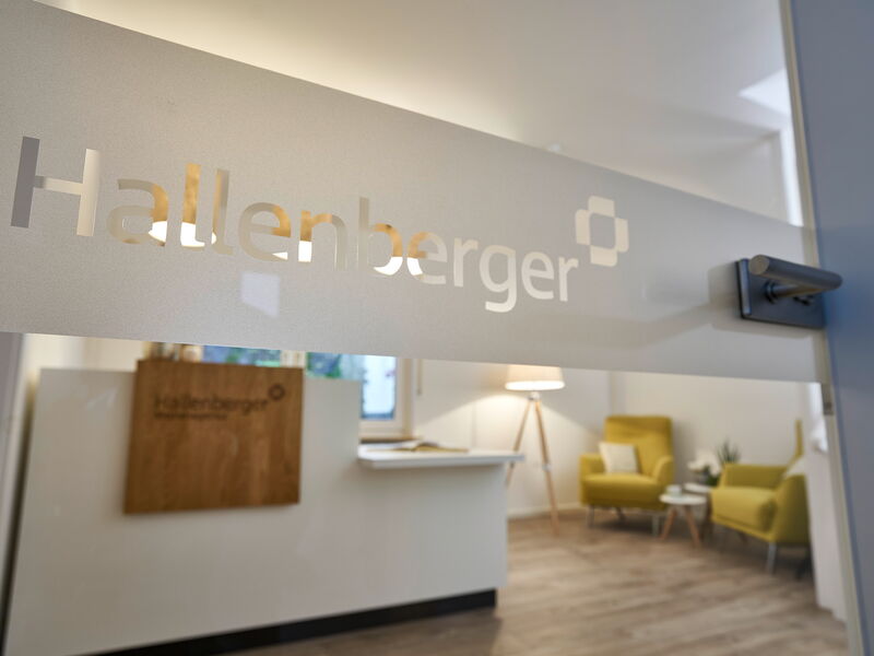 Eine Glastür mit einem Folienaufdruck "Hallenberger", durch die Tür sieht man verschwommen einen Schreibtisch, zwei grüne Sessel und eine Stehlampe.