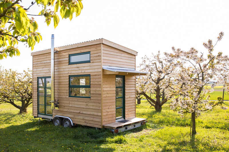 ein Tiny House mit Rädern und Schornstein der Tischlerei Bock steht auf einer Wiese umgeben von Bäumen mit blühenden Knospen
