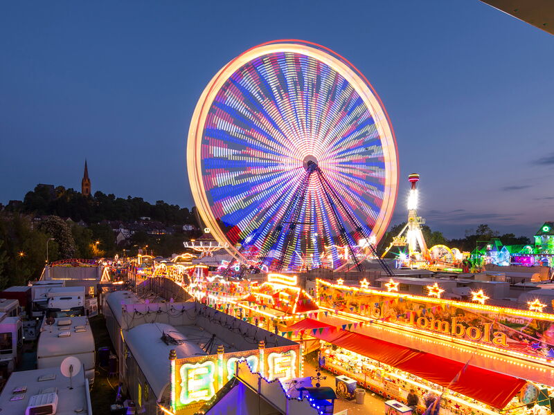 Ein großes leuchtendes Riesenrad ist zentral im Bild, welches auf dem Pfingstmarkt der Stadt Frankenberg steht und drum herum sind viele leuchtende Buden und Stände