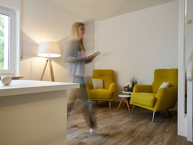 Eine blonde Frau läuft mit einem Block in der Hand durch einen Raum, im Vordergrund ein weißer Tisch, im Hintergrund zwei grüne Sessel und eine Stehlampe.