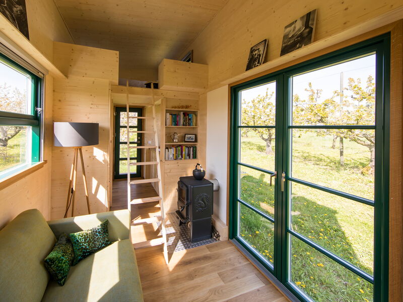 ein Tiny House der Tischlerei Bock wird von innen gezeigt mit einem Sofa, einem Ofen und einer Leiter, die hoch zum Bett führt