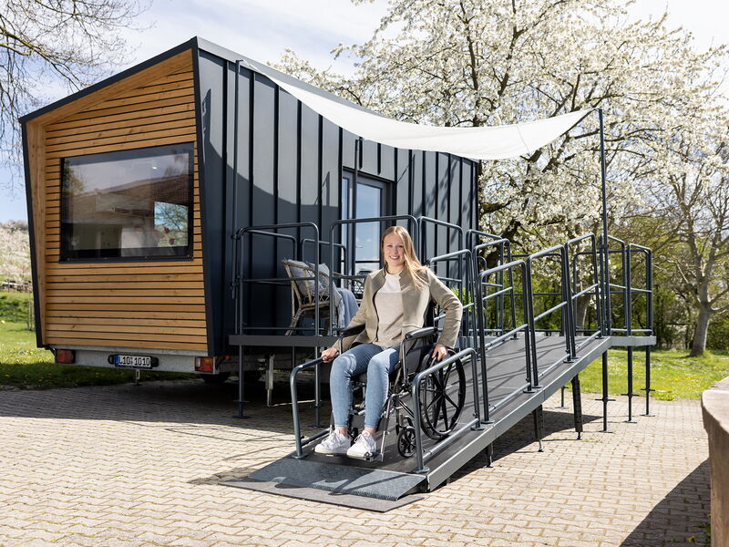 Ein barrierefreies Tiny House der Tischlerei Bock steht auf Asphalt und hat eine Rampe mit Terrasse angebaut, welche eine Rollstuhlfahrerin runterfährt