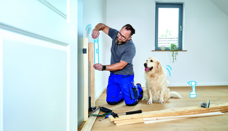 Ein Mann in Arbeitskleidung und Werkzeug kniet neben einem Hund der ihm zuschaut und misst mit einer Wasserwaage ein Brett an der Wand