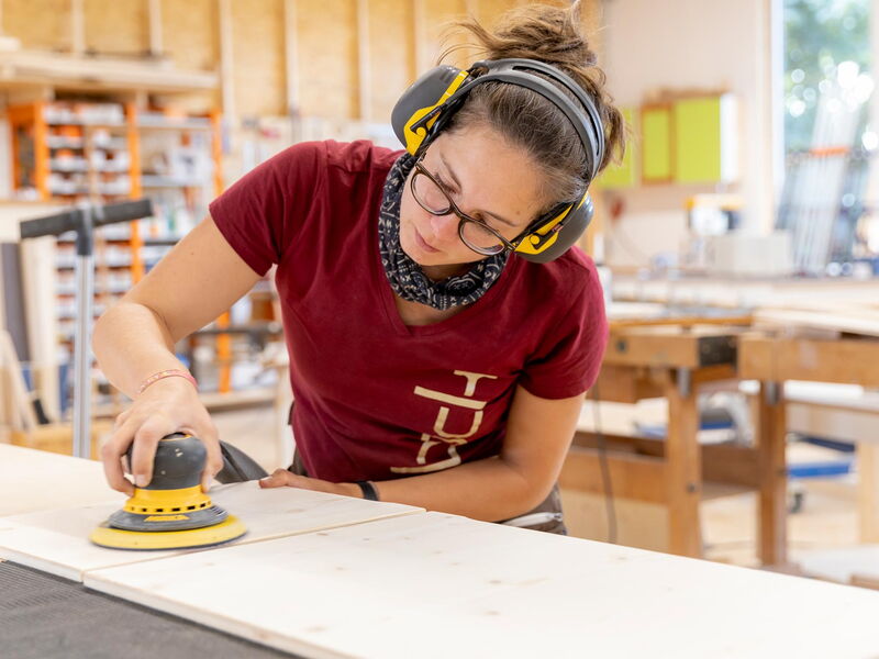 eine Schreinerin der Tischlerei Bock schleift Holz mit einer Maschine und trägt Ohrschützer