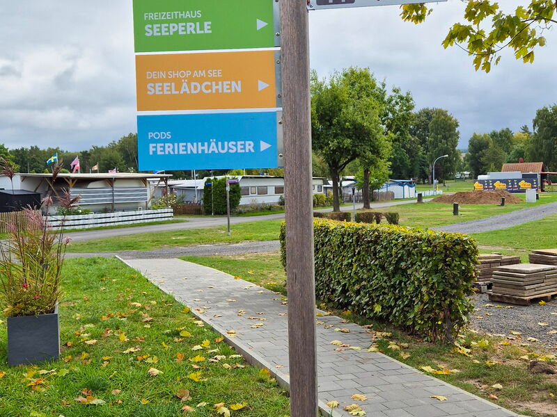 Ein Wegweiserschild auf einer Weise mit Weg zeigt mit verschiedenen Farben die Richtungen zu Saisonplätzen, dem Ferienhaus Seeperle, Seelädchen sowie den Ferienhäusern des Ferienparks Neuenhainer See.