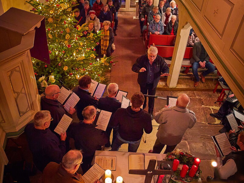 In einer Kirche mit großem Weihnachtsbaum und vielen Lichtern sitzen viele Menschen, ein Chor singt, ein beleuchteter Stern hängt an der Decke.