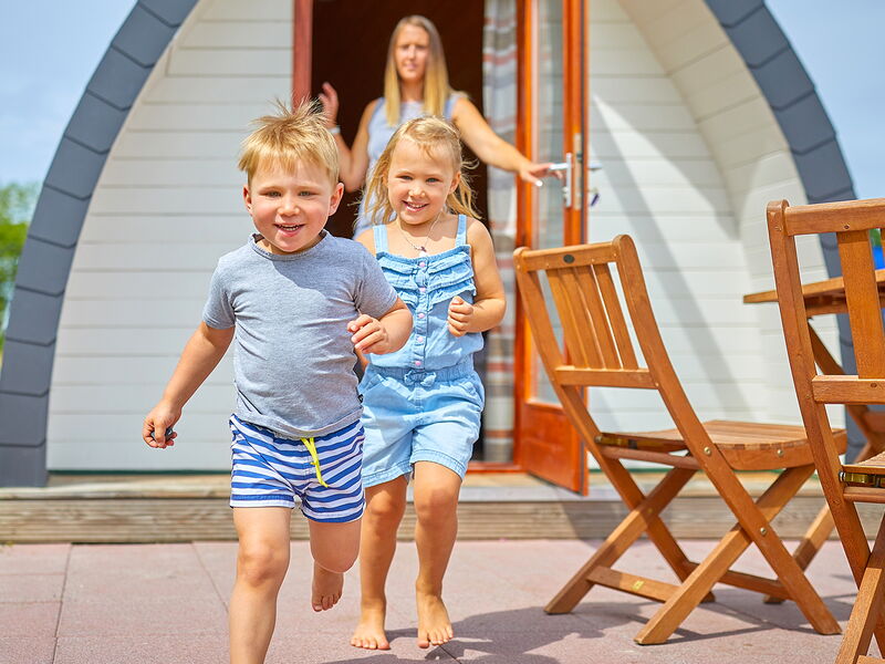 Zwei lachende Kinder rennen aus einem Holzhäuschen, die Mutter hält die Tür auf, daneben stehen Holzstühle und ein -tisch.