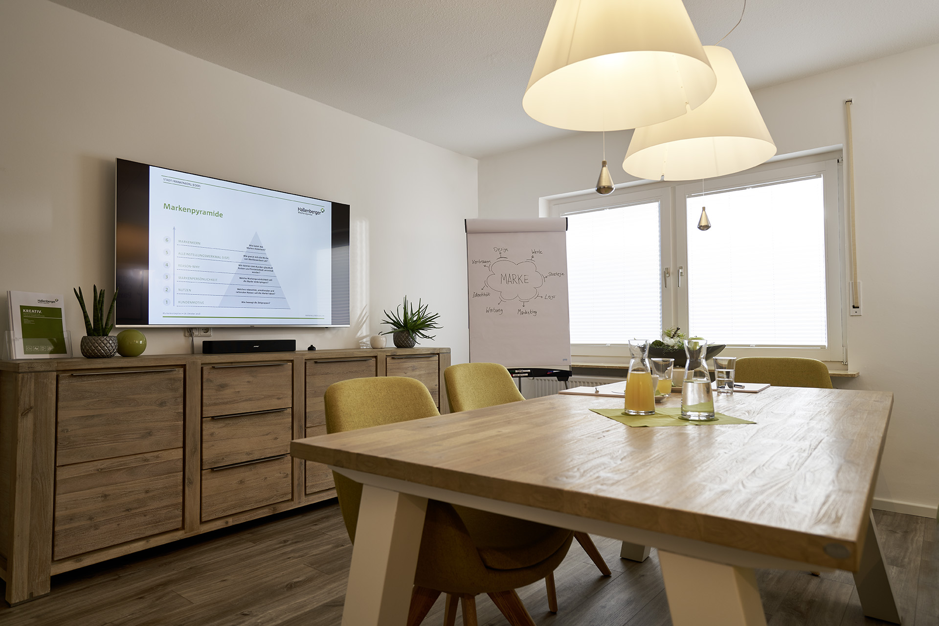 Ein Besprechungsraum mit Holztisch mit Getränken drauf, grüne Stühle drumherum, zwei Schirmlampen hängen von der Decke herunter, an der Wand hängt einer großer Bildschirm mit Bild, darunter steht ein Sideboard.