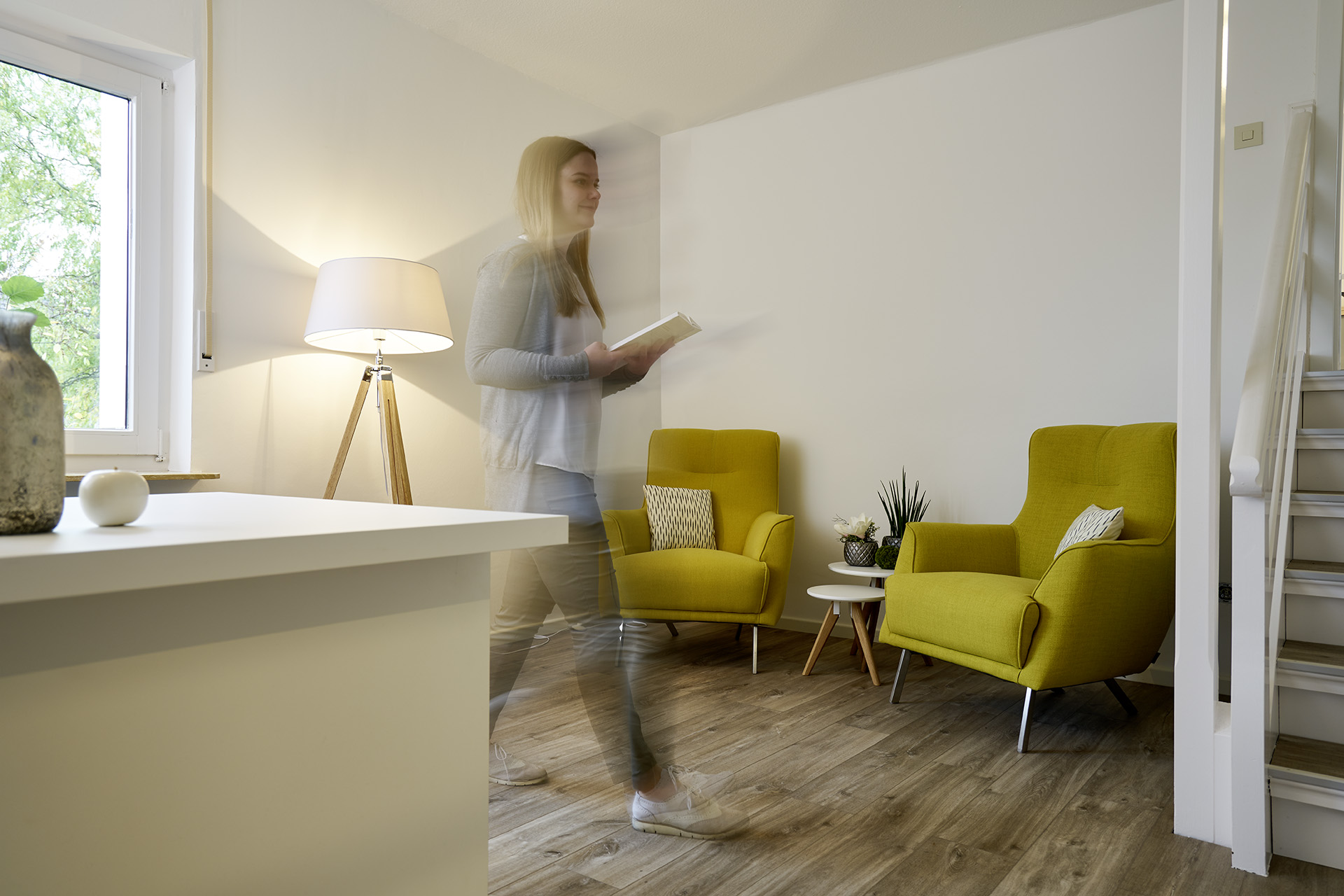 Eine blonde Frau läuft mit einem Block in der Hand durch einen Raum, im Vordergrund ein weißer Tisch, im Hintergrund zwei grüne Sessel und eine Stehlampe.