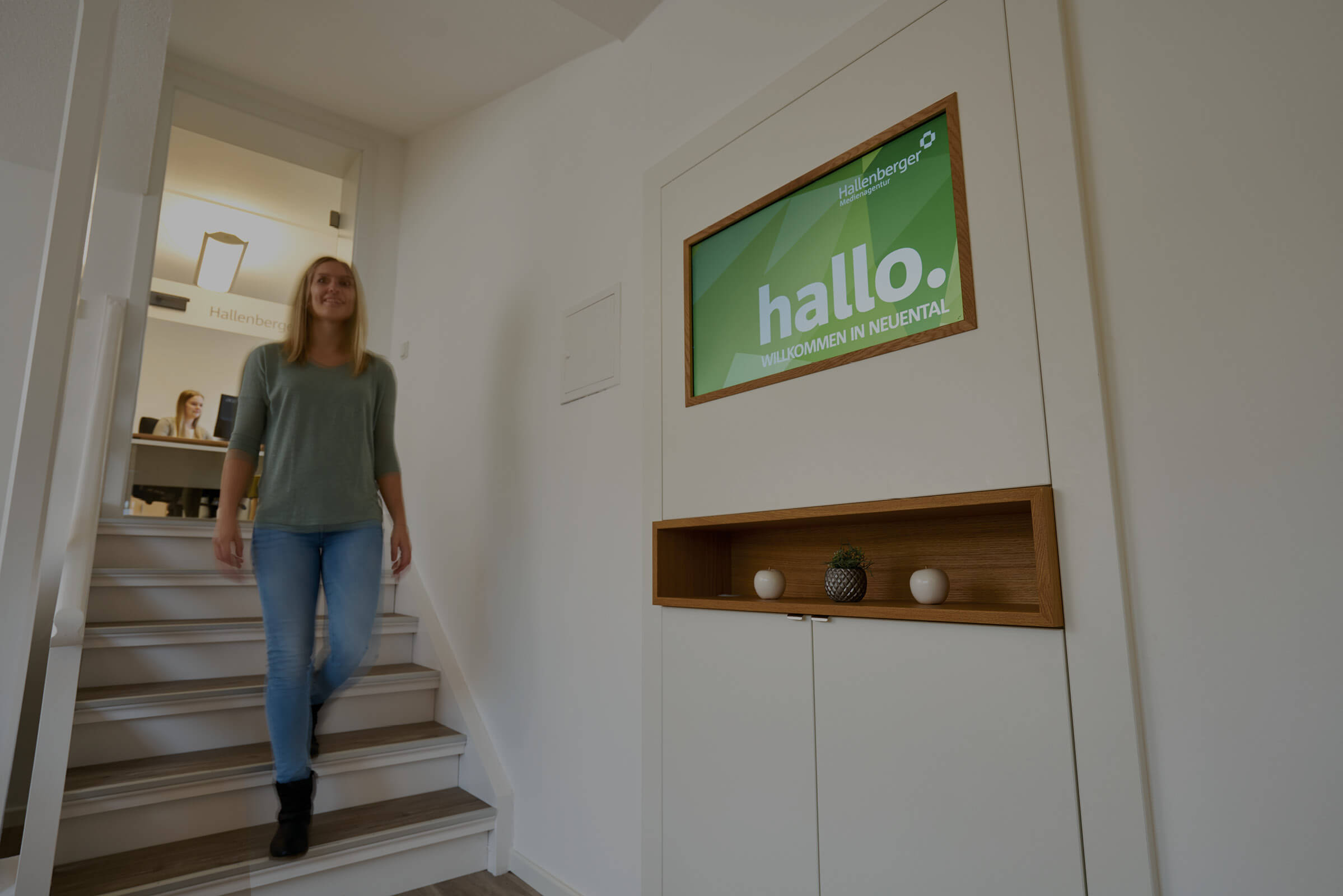 Eine Frau läuft eine Treppe hinunter, im Hintergrund ein Büroraum mit Frau am Schreibtisch, auf der rechten Seite hängt ein Bildschirm an der Wand mit einem Bild mit "hallo. Willkommen in Neuental".