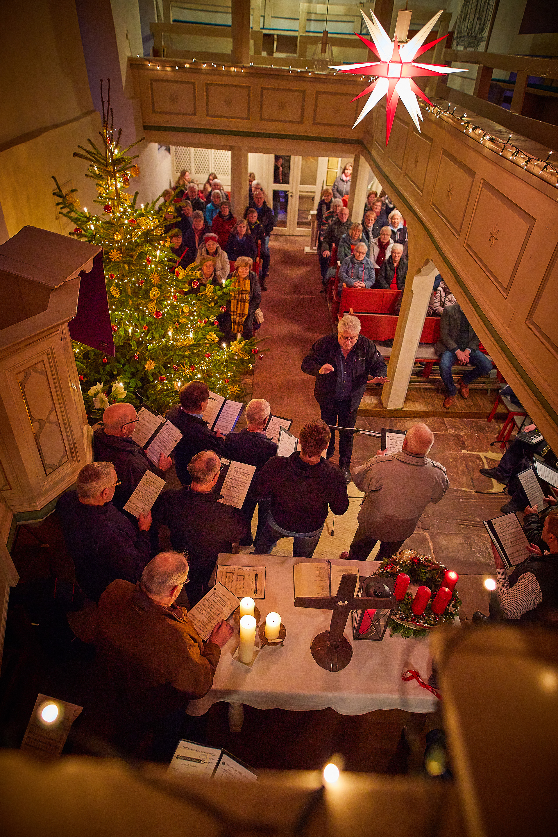 In einer Kirche mit großem Weihnachtsbaum und vielen Lichtern sitzen viele Menschen, ein Chor singt, ein beleuchteter Stern hängt an der Decke.