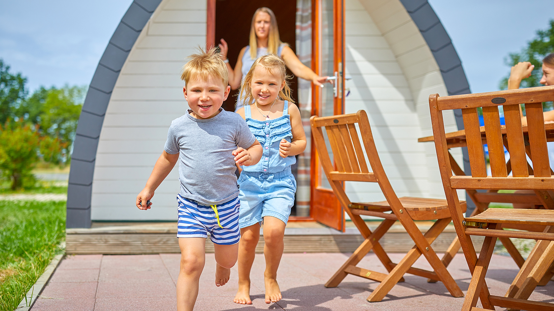 Zwei lachende Kinder rennen aus einem Holzhäuschen, die Mutter hält die Tür auf, daneben stehen Holzstühle und ein -tisch.