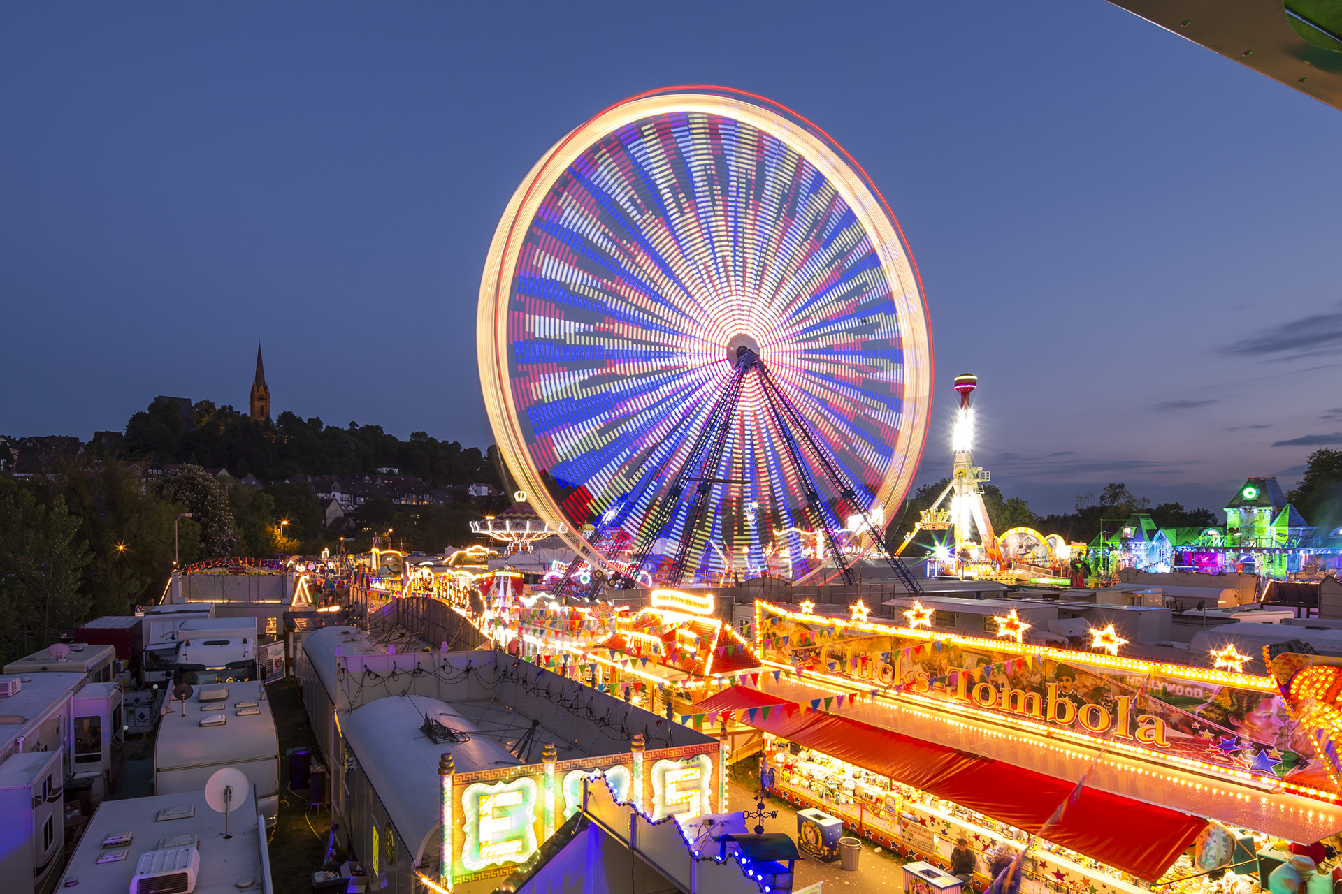 Ein großes leuchtendes Riesenrad ist zentral im Bild, welches auf dem Pfingstmarkt der Stadt Frankenberg steht und drum herum sind viele leuchtende Buden und Stände